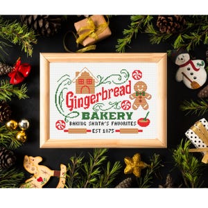 Op de afbeelding: Een ingelijst kruissteek borduurwerk met een peperkoekhuisje, een peperkoekmannetje, pepermuntjes en een deegroller. De tekst luidt Gingerbread BAKERY BAKING SANTA'S FAVORITES EST 1875 in rood en groen op een witte achtergrond.
