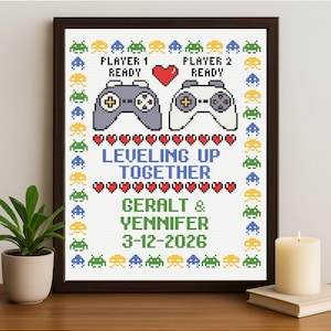 以下が含まれることがあります： 額縁に入ったクロスステッチのアートワーク。2つのゲームコントローラー、ハート、そして「LEVELING UP TOGETHER」という文字と名前と日付が表示されています。ボーダーはピクセル化されたエイリアンキャラクターで飾られています。