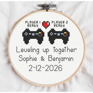 May include: Cross-stitch hoop art with a gaming theme. Features two pixelated black game controllers, a red heart, and the text "Player 1 Ready" and "Player 2 Ready". Below, it reads "Leveling up together. Sophie & Benjamin 2-12-2026".