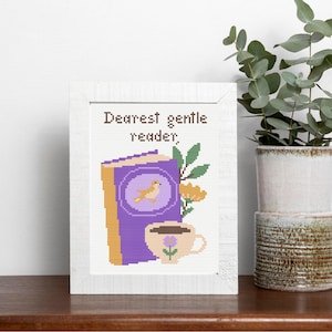 May include: A framed cross-stitch artwork featuring a purple book with a bird illustration, a cup of coffee, and the text "Dearest gentle reader." The artwork is in a white frame, with a plant in a ceramic pot in the background.