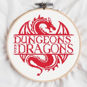 Puede incluir: Un bordado de punto de cruz en un aro de madera. El diseño presenta un dragón rojo y las palabras "DUNGEONS AND DRAGONS" en una fuente estilizada. El fondo es blanco y el aro se asegura con un cierre metálico.