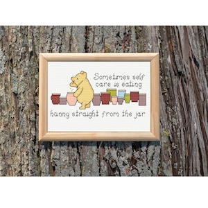 May include: A framed cross-stitch embroidery featuring a yellow bear character standing beside several colorful jars. The text reads: Sometimes self care is eating hunny straight from the jar. The piece is enclosed in a light-colored wooden frame.