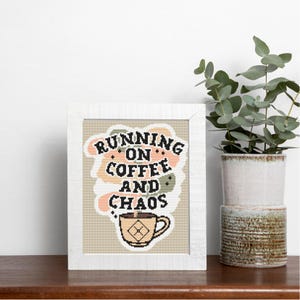 May include: A framed cross-stitch artwork with the text "RUNNING ON COFFEE AND CHAOS." The artwork features a coffee cup graphic and a background of peach, green, and beige colors. The frame is white, and the artwork is displayed on a wooden surface.