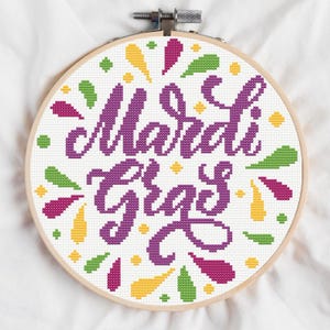 May include: A circular cross-stitch embroidery featuring the words "Mardi Gras" in purple. The design incorporates colourful accents in yellow, green, and pink, set against a white backdrop. The wooden hoop is secured with a metal clasp.
