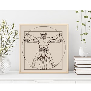 May include: Framed cross-stitch art featuring a muscular figure in a circle and square, reminiscent of Da Vinci's Vitruvian Man. The artwork is in shades of brown and beige, with a gold-colored frame. The piece is displayed on a white surface.