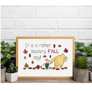Könnte beinhalten: Ein gerahmtes Kreuzstichbild mit Winnie Puuh und Ferkel in einer Herbstszene. Der Text lautet "It is a rather blustery FALL day!" mit Herbstblättern. Der Rahmen ist aus hellem Holz.
