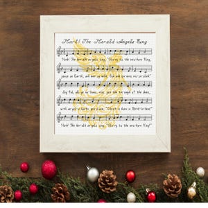 May include: A square framed cross-stitch piece featuring the musical notation and lyrics for Hark! The Herald Angels Sing. A large, golden, stylized angel silhouette is embroidered behind the black musical staff and text on white fabric.