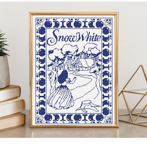 May include: A framed cross-stitch pattern in blue and white featuring the text Snow White. The design depicts a figure in a long skirt standing on a path leading to a cottage, surrounded by trees and a border of apple motifs in a gold frame.