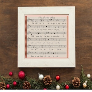 May include: A framed cross-stitch embroidery featuring the musical notation and lyrics for the Christmas carol The First Noel. The piece is set in a white wooden frame and displays black musical notes and text on a light fabric background.