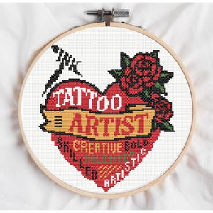 Puede incluir: Bordado de punto de cruz en un aro de madera. El diseño presenta un corazón rojo con las palabras "TATTOO ARTIST" y una pancarta que dice "SKILLED CREATIVE BOLD ARTISTIC". Rosas rojas y un tintero negro completan el diseño.