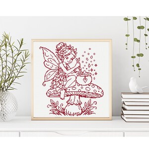 May include: A framed cross-stitch pattern featuring a dark red line art illustration of a fairy with wings sitting on a large spotted mushroom. The fairy is stirring a small cauldron with a heart symbol, surrounded by small sparkling star shapes.