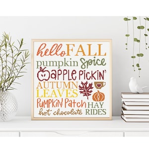 Op de afbeelding: Een ingelijst kruissteekkunstwerk met de woorden "hello FALL" in oranje, "pumpkin Spice" in groen en "apple Pickin'" in rood. Andere woorden zijn "AUTUMN LEAVES", "PUMPKIN Patch", "hot chocolate RIDES".