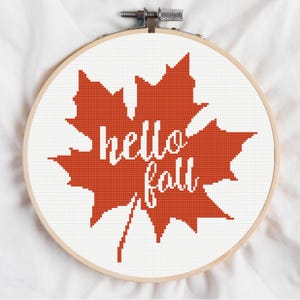 May include: A cross-stitch embroidery hoop with an orange maple leaf design. The words "hello fall" are stitched in white within the leaf. The embroidery is set in a wooden hoop against a white fabric background.
