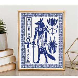 May include: A framed cross-stitch pattern featuring a dark blue depiction of the Egyptian deity Anubis, an ankh symbol, hieroglyphics, and a lotus flower. The design is enclosed in a geometric Greek key border, all set against a white background.