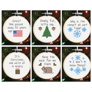 Puede incluir: Seis adornos navideños bordados en aros de madera. Cada uno presenta una cita humorística en punto de cruz, con imágenes festivas como un árbol de Navidad, copos de nieve y un pavo asado. Los adornos están colocados sobre un fondo de luces navideñas.