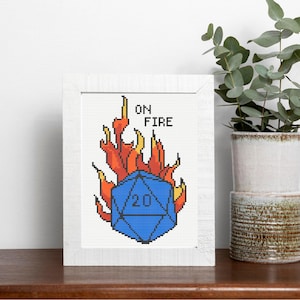 May include: A framed cross-stitch depicting a blue twenty-sided die in flames. The words "ON FIRE" are stitched above the die. The artwork is set against a white background within a white frame.