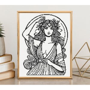 May include: A framed cross-stitch pattern featuring a black and white line art illustration of a woman with long flowing hair, a floral headpiece, and a crescent moon symbol on her forehead, set against a circular background within a gold frame.