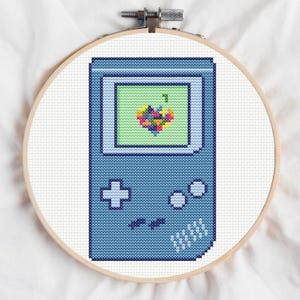 Può includere: Ricamo a punto croce di una console di videogiochi portatile retrò in un telaio di legno. La console è blu con uno schermo verde che mostra un design pixelato colorato. Il punto croce è ambientato su uno sfondo di tessuto bianco.