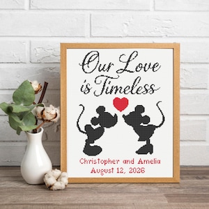 以下が含まれることがあります： 黒い文字でOur Love is Timeless、赤いハート、2つの黒いネズミのシルエットが描かれた額入りのクロスステッチパターン。シルエットの下には、赤い文字でChristopher and Amelia、その下に2026年8月12日という日付が記されています。