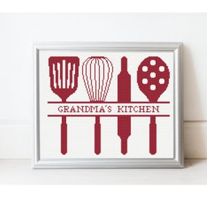 May include: A framed cross-stitch featuring kitchen utensils: a spatula, whisk, rolling pin, and masher. The text "GRANDMA'S KITCHEN" is stitched below the utensils in a red hue. The frame is silver.