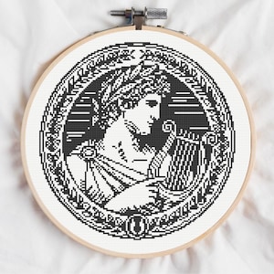 May include: A circular wooden embroidery hoop containing a black and white cross-stitch pattern. The design features a profile of a figure wearing a laurel wreath and playing a lyre, enclosed within a decorative circular border of geometric patterns.
