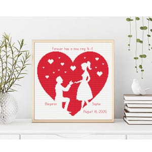 May include: A framed cross-stitch artwork featuring a red heart with a couple in silhouette. The man is kneeling, proposing to the woman. The text reads "Forever has a nice ring to it," with names and a date below. The frame is gold.