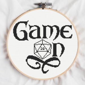 Może przedstawiać: Obręcz do haftu krzyżykowego z czarnymi słowami "Game On" i grafiką kości. Wzór znajduje się na białej tkaninie, w drewnianej obręczy. Tekst i grafika są pogrubione i stylizowane.