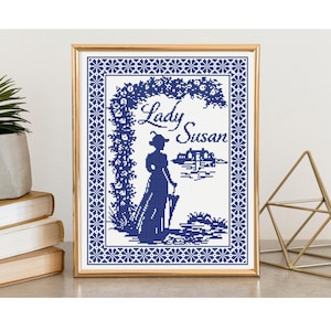 May include: A framed cross-stitch pattern featuring a dark blue silhouette of a person in a long dress holding an umbrella under a floral archway. The text Lady Susan is written in script above the figure, with a house in the background.