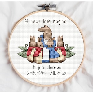 Può includere: Ricamo a punto croce in un telaio di legno. Il disegno presenta quattro coniglietti, due con giacche rosse e due in blu. Il testo recita "A new tale begins." e include il nome "Elijah James" e i dettagli della nascita.