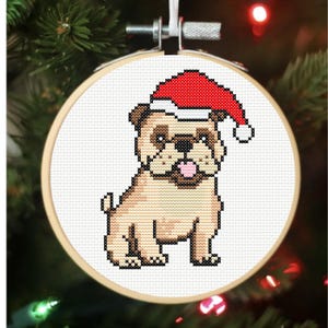 May include: A cross-stitch embroidery hoop featuring a pug wearing a Santa hat. The pug is tan, brown, and black, with a pink tongue. The Santa hat is red with a white trim and a white pom-pom. The hoop is made of wood and is hanging on a Christmas tree.
