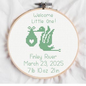 May include: A cross-stitch embroidery hoop with the text "Welcome Little One!" and a stork carrying a baby. The text "Finley River, March 23, 2025, 7lb 10oz 21in" is also included. The design is in green on a white background.