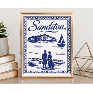 May include: A framed cross-stitch artwork featuring a blue and white design with the word "Sanditon" at the top. The artwork depicts a seaside scene with a building, sailboat, and figures. The frame is gold.