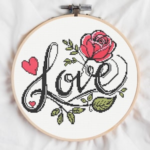 May include: A circular wooden embroidery hoop containing a cross-stitch pattern on white fabric. The design features the word LOVE in black script lettering, accented by a pink rose with green leaves and two small pink hearts.