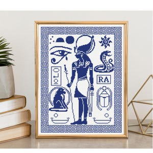 Puede incluir: Un patrón de punto de cruz enmarcado con un diseño egipcio azul oscuro sobre fondo blanco. La obra muestra una figura central, el Ojo de Horus, una cobra, un escarabajo, un babuino sentado y el texto RA, todo dentro de un borde de greca.