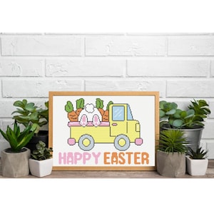 Puede incluir: Un diseño de punto de cruz enmarcado con un camión amarillo que transporta zanahorias y un conejito. El texto "HAPPY EASTER" se muestra en rosa y naranja. El diseño está sobre una pared de ladrillos blancos, con plantas en macetas en primer plano.