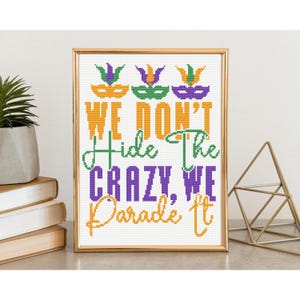 Könnte beinhalten: Gerahmtes Kreuzstich-Kunstwerk in einem goldenen Rahmen. Das Kunstwerk zeigt den Text "WE DON'T HIDE THE CRAZY, WE Parade It" in Orange, Grün und Lila. Über dem Text befinden sich drei Maskenmotive in den gleichen Farben. Das Kunstwerk wird auf einem Tisch ausgestellt.