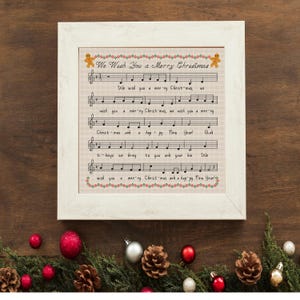 May include: A framed cross-stitch piece featuring the musical notation and lyrics for We Wish You a Merry Christmas. The design includes gingerbread men, a red and green border, and black text on a light fabric background in a white frame.