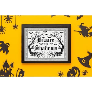 May include: A black framed cross stitch piece featuring the text "Beware the Shadows" in a gothic font, surrounded by black silhouettes of bare trees, small bats, and a decorative border on a white fabric background within the frame.