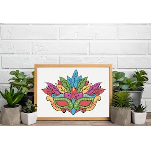 May include: A framed cross-stitch of a colorful carnival mask. The mask features a vibrant design with red, green, yellow, blue, and purple feathers. The mask is set against a white background and framed in a light brown wooden frame.