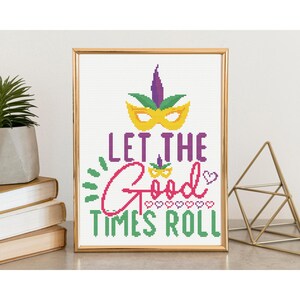May include: A framed cross-stitch artwork with a gold frame. The design features a yellow mask, purple feathers, and the phrase "LET THE GOOD TIMES ROLL" in green and pink. The artwork is displayed on a white surface.