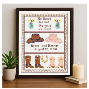 Könnte beinhalten: Gerahmtes Kreuzstich-Kunstwerk mit braunem Rahmen. Das Design umfasst einen braunen Cowboyhut, einen rosa Hut, Cowboy-Stiefel und ein Hufeisen. Der Text lautet: "He tipped his hat, She gave her heart. Robert and Hannah August 12, 2026."