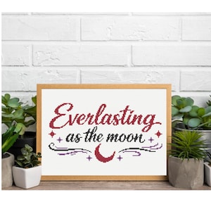 May include: A framed cross-stitch piece with the words "Everlasting as the moon" in burgundy script. The design includes a crescent moon and stars, set against a white background. The frame is a light wood color.