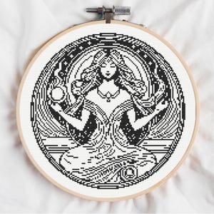 May include: A circular wooden embroidery hoop containing a cross-stitch pattern of a figure with long hair, seated in a meditative pose. The design is rendered in black thread on white fabric, featuring intricate geometric details and a circular border.