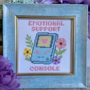 May include: Cross-stitch art in a light blue wooden frame. The artwork features a light blue handheld console with a pink heart on the screen, surrounded by colorful flowers. The words "EMOTIONAL SUPPORT CONSOLE" are stitched in pink.