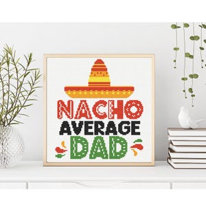 May include: Framed cross-stitch art with the text "NACHO AVERAGE DAD" in red, black, and green. A yellow and orange sombrero sits above the text. The artwork is displayed on a white surface.