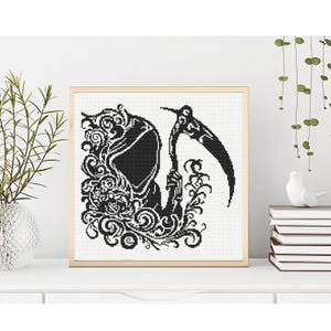 May include: A square cross stitch pattern featuring a black silhouette of a hooded grim reaper figure holding a scythe, surrounded by intricate swirling floral and vine motifs, all presented within a light-colored wooden frame.