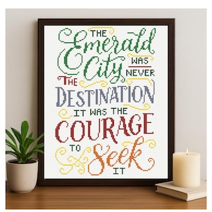 Puede incluir: Una obra de arte en punto de cruz enmarcada con la cita "The Emerald City Was Never The Destination, It Was The Courage To Seek It." El texto es en verde, azul y naranja sobre un fondo blanco. Se ven una pequeña planta en maceta y una vela.
