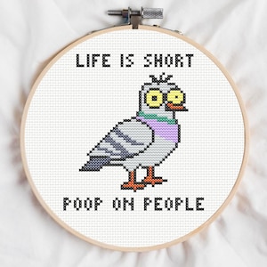 May include: A circular wooden embroidery hoop containing white fabric with a cross-stitch design of a grey pigeon with yellow eyes and a purple neck. The text above the bird reads LIFE IS SHORT and the text below reads POOP ON PEOPLE.