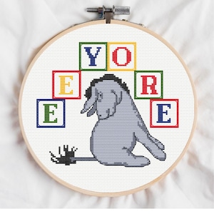 Könnte beinhalten: Ein Kreuzstich-Stickrahmen mit Eeyore aus Winnie Puuh. Der graue Esel ist mit einem traurigen Ausdruck gestickt. Das Wort "Eeyore" ist in bunten Blockbuchstaben geschrieben. Der Rahmen ist aus Holz.