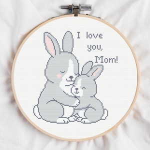 May include: Cross-stitch embroidery hoop featuring two gray bunnies hugging, with the text "I love you, Mom!" in a playful font. The bunnies have pink accents on their ears and cheeks, set against a white background.